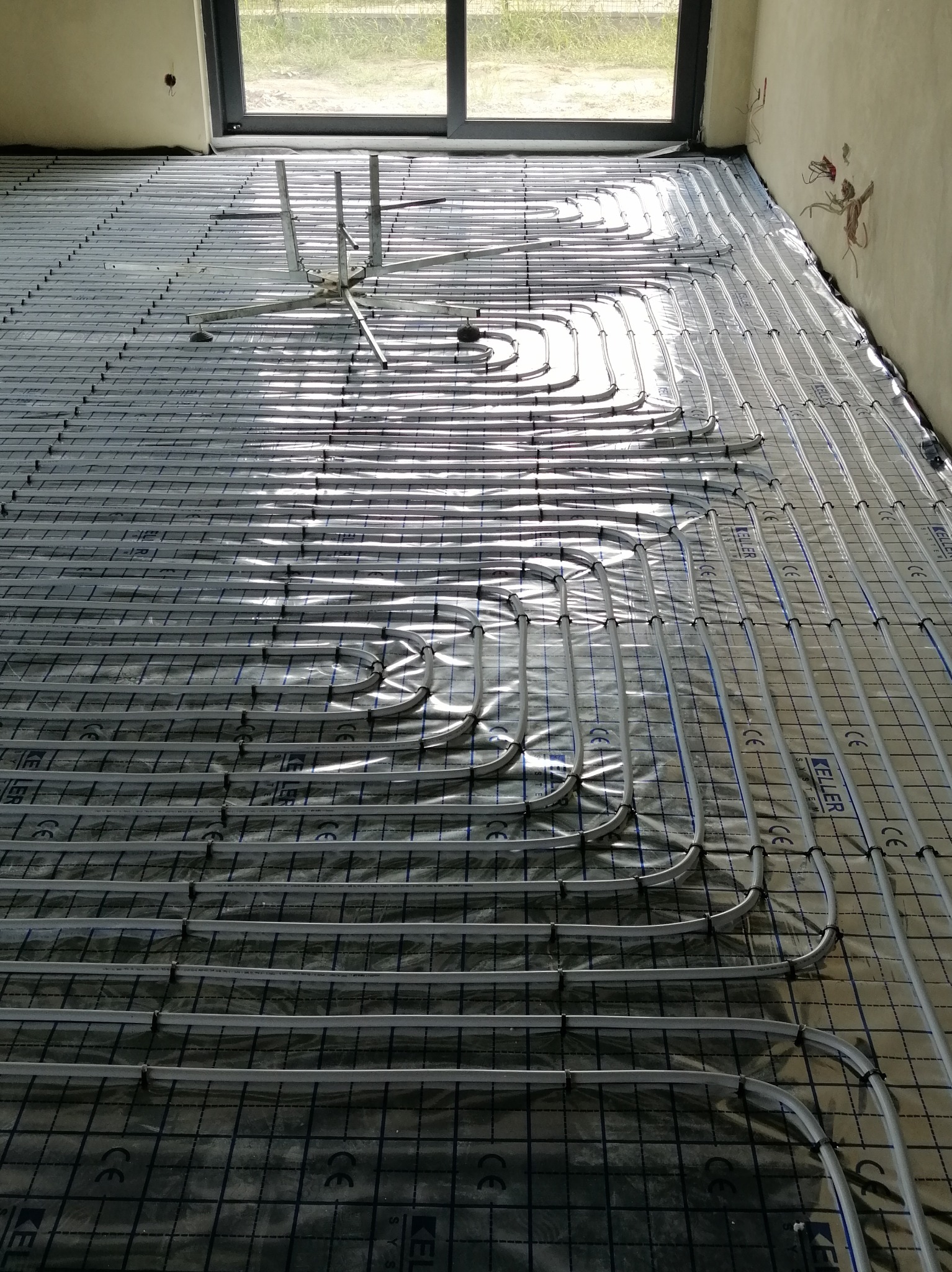 Rozłożone rury ogrzewania podłogowego na folii z nadrukiem, widoczne mocowania rur, w tle okno i surowa ściana.