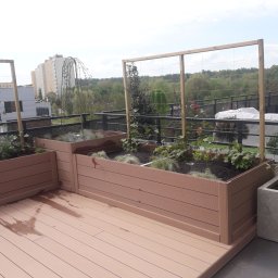 Adam Kuszczyk GC usługi ogrodnicze - Zaprojektowany taras na dachu z drewnianą podłogą i podwyższonymi rabatami z roślinami, widok na miasto i zieleń w tle. Elementy drewniane tworzą kraty dla pnączy.
