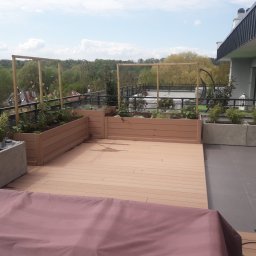 Adam Kuszczyk GC usługi ogrodnicze - Umeblowany taras na dachu z drewnianą podłogą i podwyższonymi grządkami z roślinami, widok na zieleń w tle, słoneczny dzień.