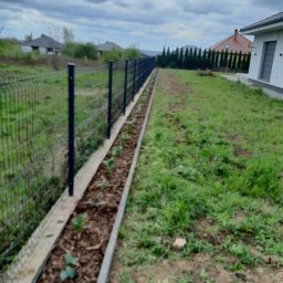 Adam Kuszczyk GC usługi ogrodnicze - Rabata z młodymi sadzonkami wzdłuż ogrodzenia panelowego na tle domu i pochmurnego nieba. Widok na założony ogród w trakcie realizacji.