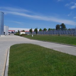 Adam Kuszczyk GC usługi ogrodnicze - Widok na teren przemysłowy z panelami słonecznymi za ogrodzeniem, zbiornikiem i budynkiem w tle, w słoneczny dzień. Fragment samochodu dostawczego po lewej.