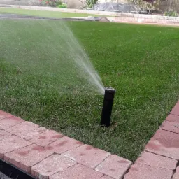 Automatyczny system nawadniania trawnika w ogrodzie, zraszacz w akcji, widoczna woda pod ciśnieniem, obrzeże z czerwonej kostki.