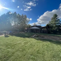 Układanie trawy z rolki w ogrodzie, widoczni pracownicy w trakcie pracy, nasłoneczniony kadr z błękitnym niebem i białymi chmurami w tle.