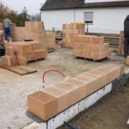 Power-Dial - Budowa ściany z pustaków ceramicznych na fundamencie, widoczne zapasy materiału i narzędzia na placu budowy, częściowo w pochmurny dzień.