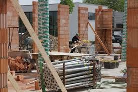 Wznoszenie ścian z cegły na placu budowy, widoczne podpory i narzędzia, pracownik w tle, częściowo ukończona konstrukcja.