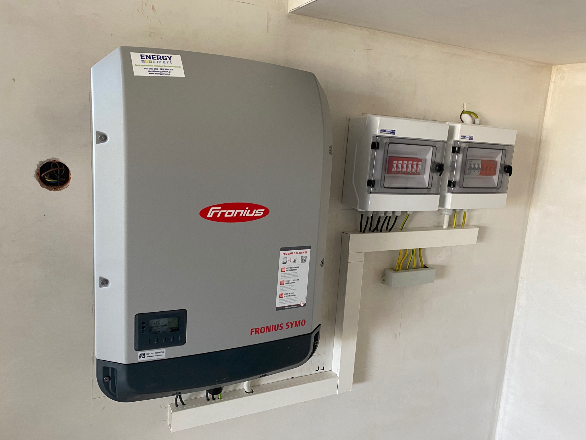 Szary inwerter Fronius Symo zamontowany na ścianie obok dwóch białych skrzynek rozdzielczych z przewodami. Widoczne naklejki informacyjne na inwerterze i logo firmy Energy Smart.