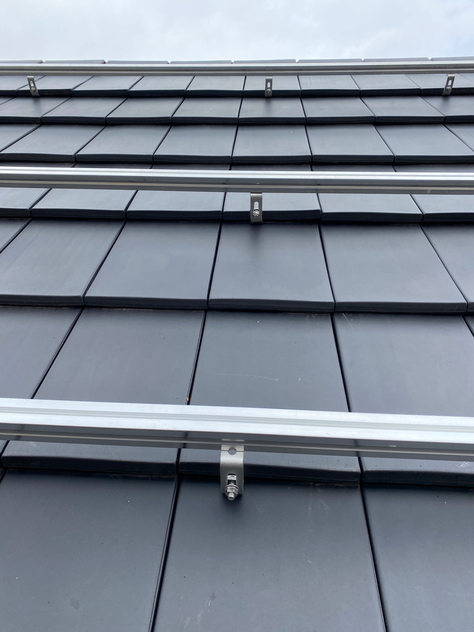 Montaż szyn aluminiowych do mocowania paneli na dachu pokrytym czarną dachówką ceramiczną, widoczne metalowe uchwyty i śruby mocujące.