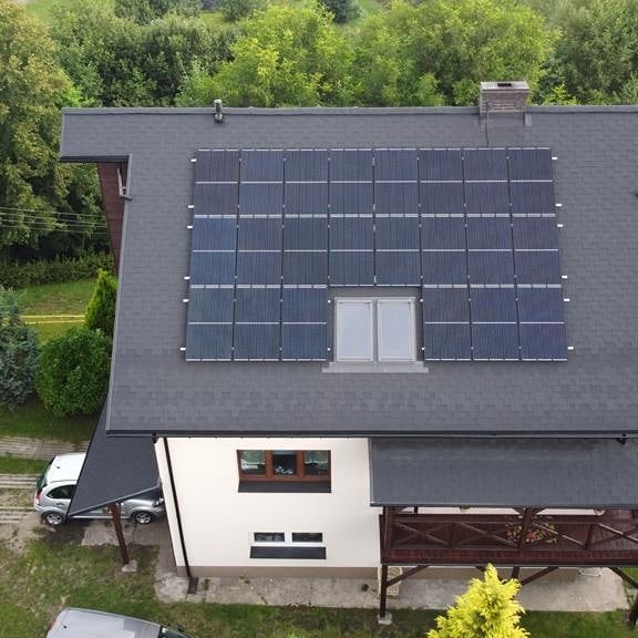 Panele słoneczne zamontowane na dachu domu jednorodzinnego ze spadzistym dachem pokrytym szarą dachówką bitumiczną. Moduły fotowoltaiczne umieszczone nad oknem dachowym. Widok z góry na dom...