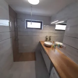 Nowoczesna łazienka z drewnianym blatem, owalną umywalką i kabiną prysznicową typu walk-in, oświetlona lampą sufitową i listwą LED nad lustrem.