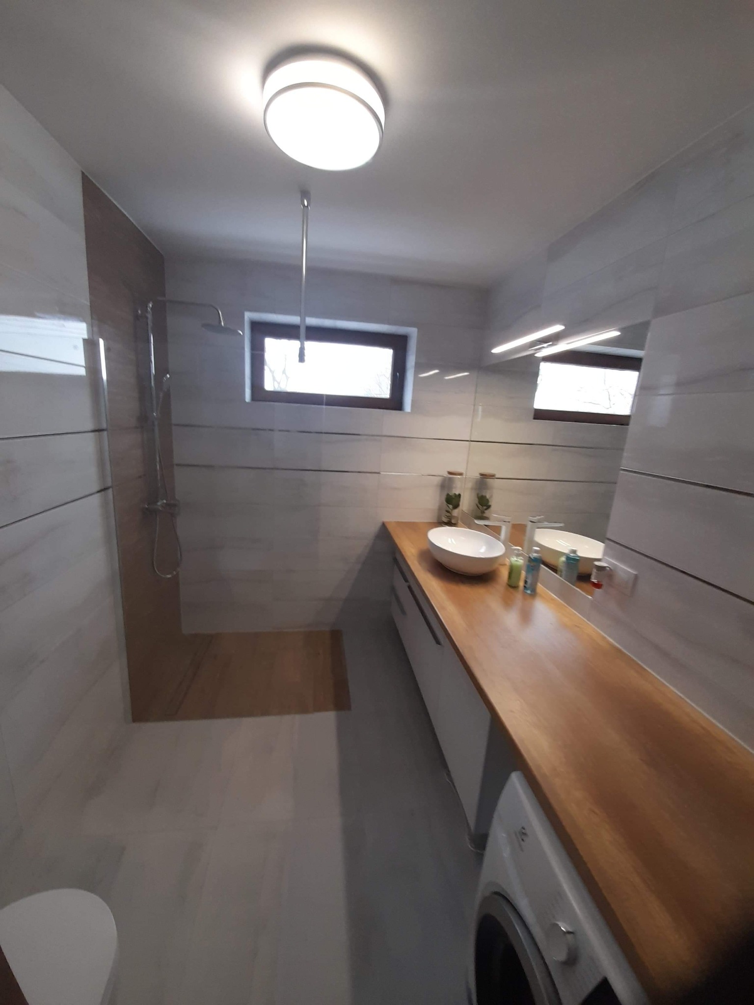 Nowoczesna łazienka z drewnianym blatem, owalną umywalką i kabiną prysznicową typu walk-in, oświetlona lampą sufitową i listwą LED nad lustrem.