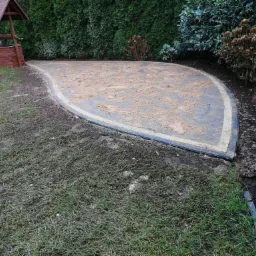 Niedawno ułożona, łukowata powierzchnia z kostki brukowej w Kartuzach, pokryta piaskiem, z widoczną studnią zadaszoną drewnianą konstrukcją w tle.