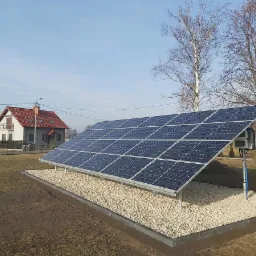 Panel słoneczny na żwirowym podłożu, w tle dom z czerwonym dachem i drzewa bez liści, błękitne niebo.