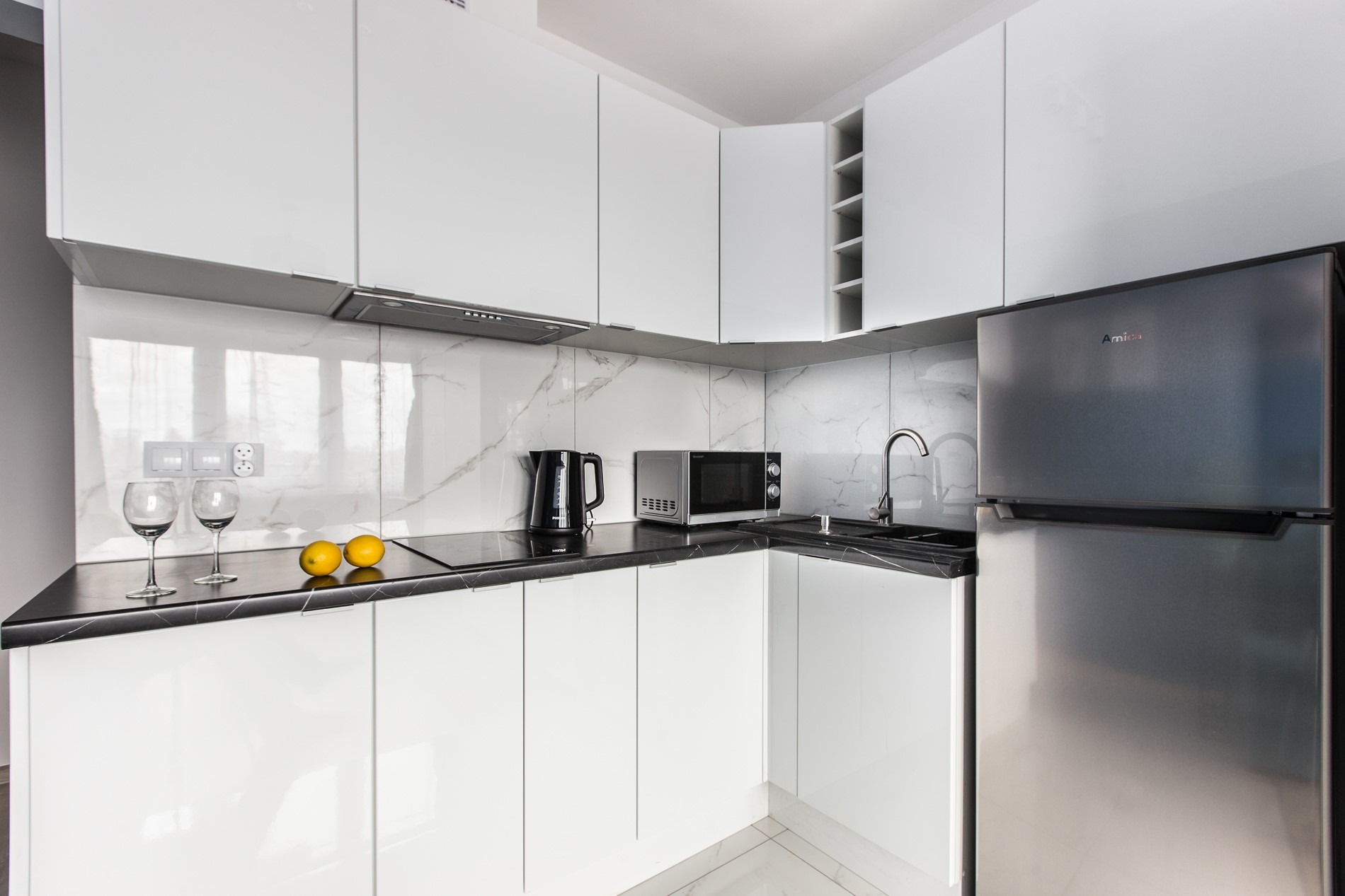 Nowoczesna, biała kuchnia z marmurowym backsplash, czarnym blatem, srebrną lodówką i sprzętami AGD; na blacie dwa kieliszki i cytryny.