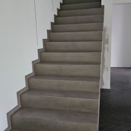 MiP RESIN - Schody wykończone szarym mikrocementem, kontrastujące z ciemną podłogą ułożoną w jodełkę. Minimalistyczne wnętrze z białymi ścianami i widocznym oknem.