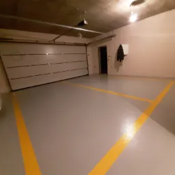 Szara, błyszcząca podłoga z żółtymi liniami w garażu w Opacz Mała, widoczna brama garażowa i wieszak z kurtką na ścianie.