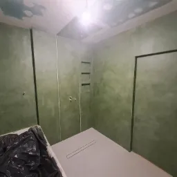 Łazienka w trakcie wykończenia z zielonymi ścianami o nieregularnej fakturze, widoczna kratka odpływowa w podłodze i pojemnik z czarną folią zabezpieczającą.