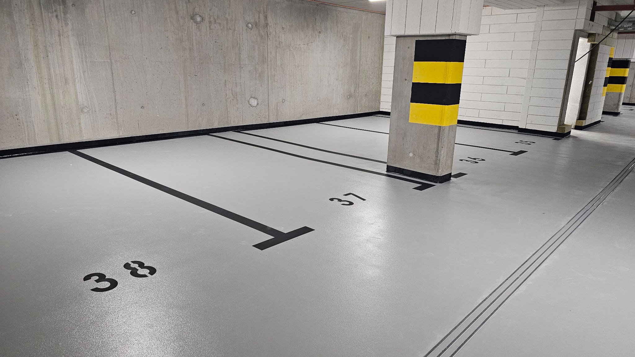 Szara posadzka w garażu podziemnym z czarnymi liniami wyznaczającymi miejsca parkingowe i numerami 38, 37, 36. Słupy w czarno-żółte pasy. Betonowa ściana w tle.