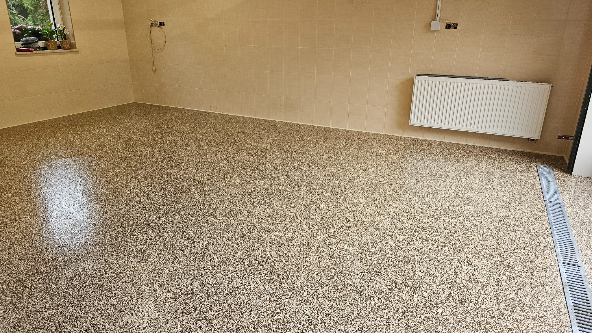 Beżowa posadzka z drobnym wzorem w pomieszczeniu z grzejnikiem i kratką odpływową. Widoczne płytki na ścianach i okno z roślinami.