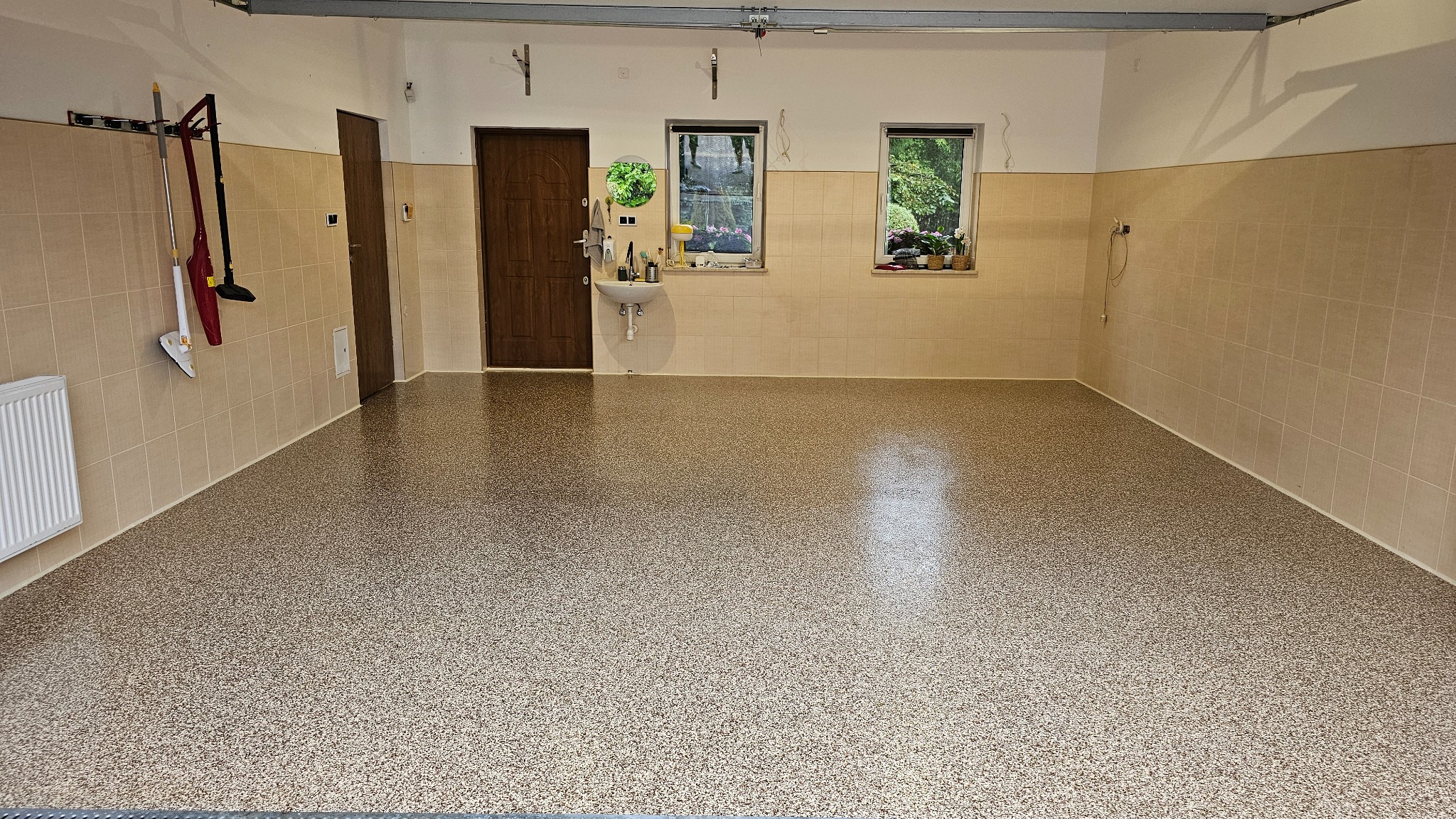 Jasna, beżowo-brązowa posadzka w garażu z kafelkowymi ścianami. Widoczne okna, drzwi, umywalka i akcesoria do sprzątania. Gładka powierzchnia, równomierne oświetlenie.