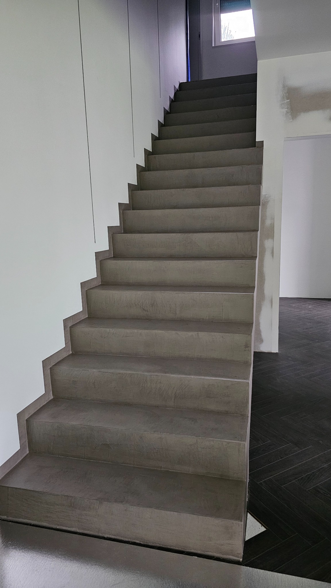 Schody wykończone szarym mikrocementem, kontrastujące z ciemną podłogą ułożoną w jodełkę. Minimalistyczne wnętrze z białymi ścianami i widocznym oknem.