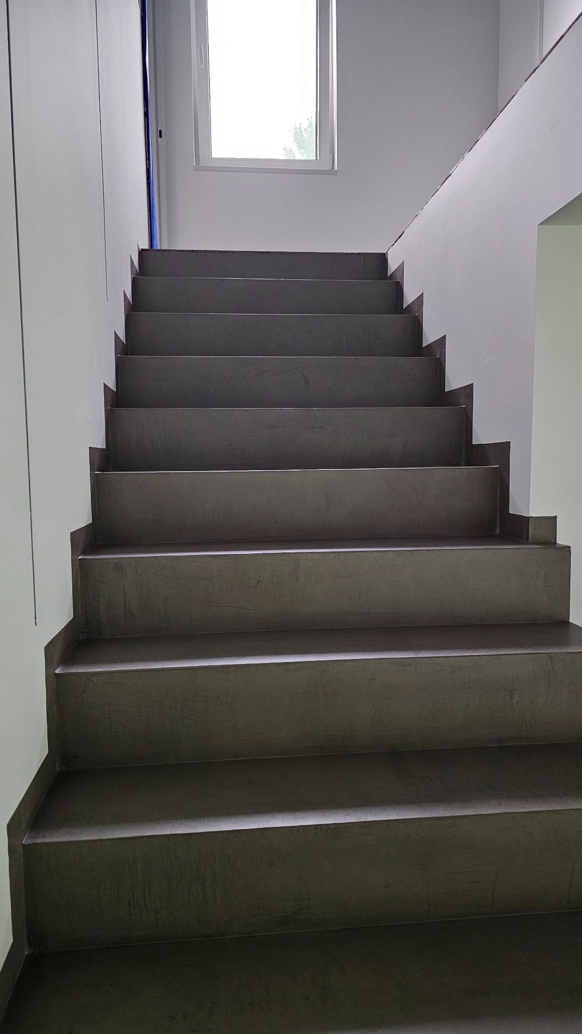 Schody wykończone szarą posadzką. Widoczne stopnie i fragment okna na górze. Minimalistyczny design, gładka powierzchnia, jednolite wykończenie.