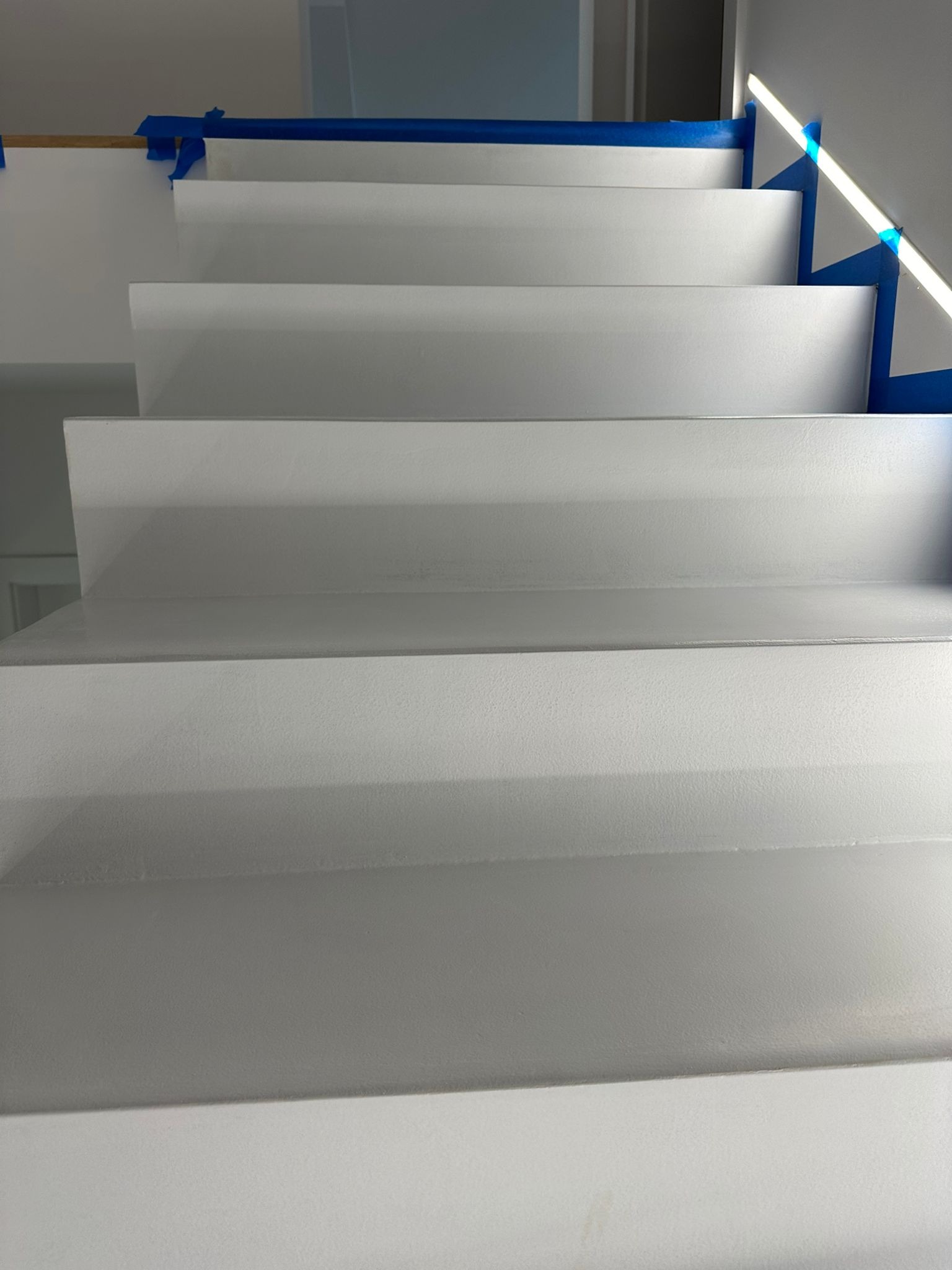 Białe schody z zabezpieczeniem z niebieskiej taśmy malarskiej, widok z dołu, świeżo po nałożeniu powłoki. Minimalistyczny design, jasne wnętrze.