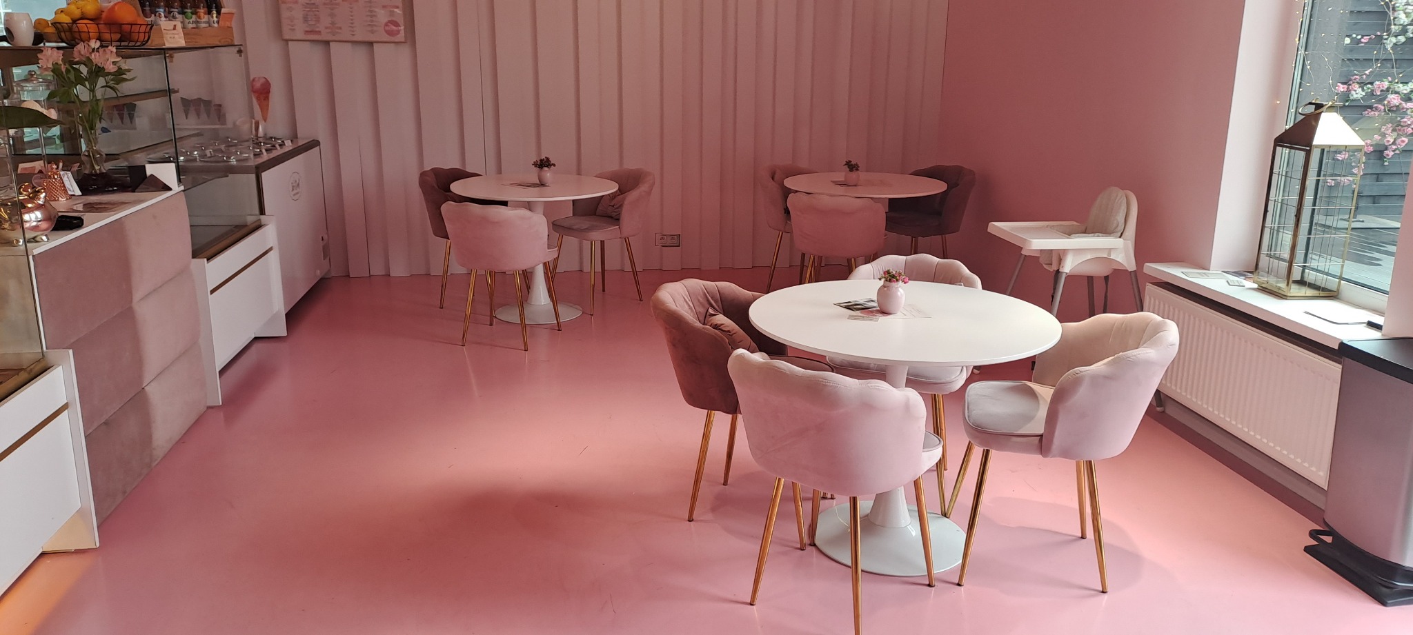 Różowa posadzka żywiczna w kawiarni z welurowymi krzesłami i białymi stolikami. Widoczny fragment lady chłodniczej i okno z dekoracją.