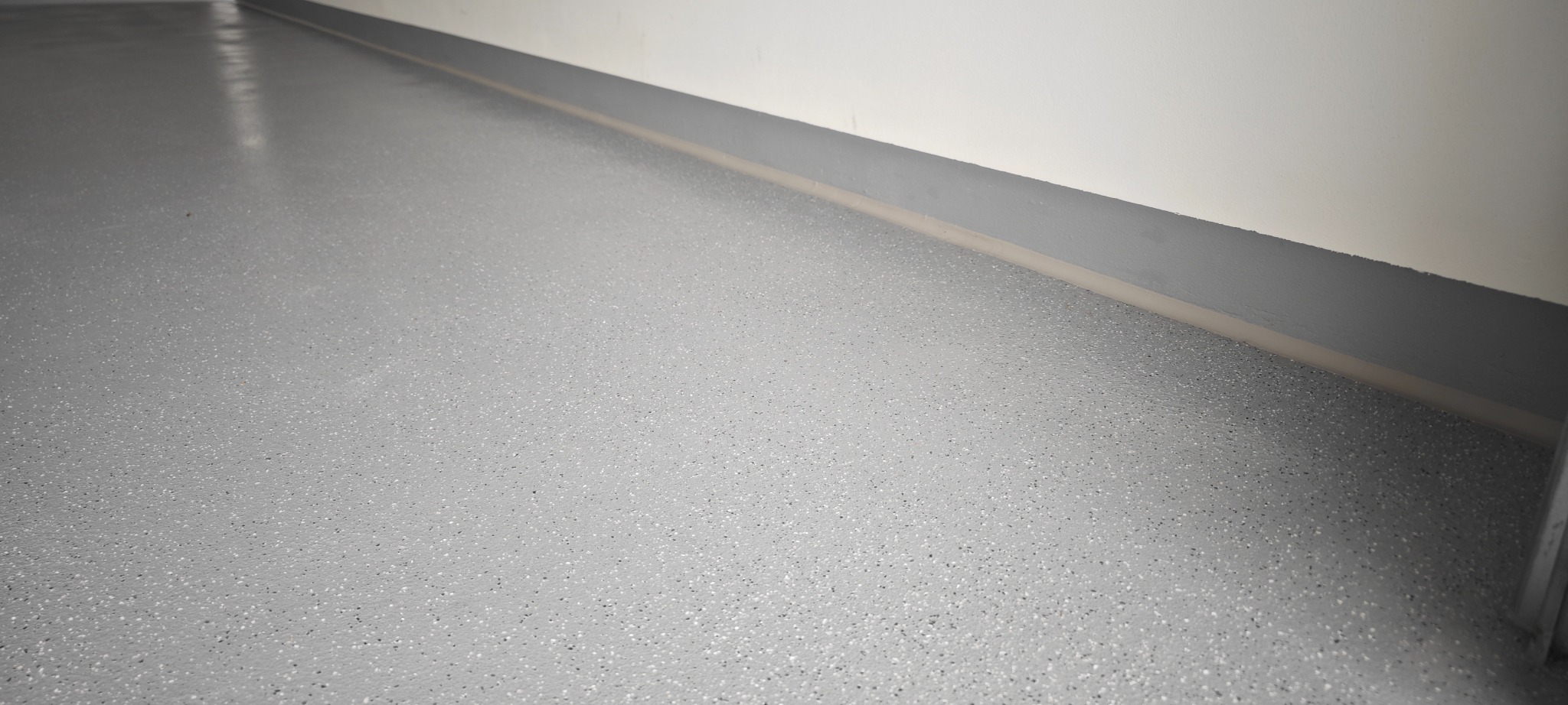 Jasnoszara, gładka posadzka z drobnymi, ciemnymi plamkami, przy ścianie z szarą listwą przypodłogową. Widoczny fragment futryny drzwi.