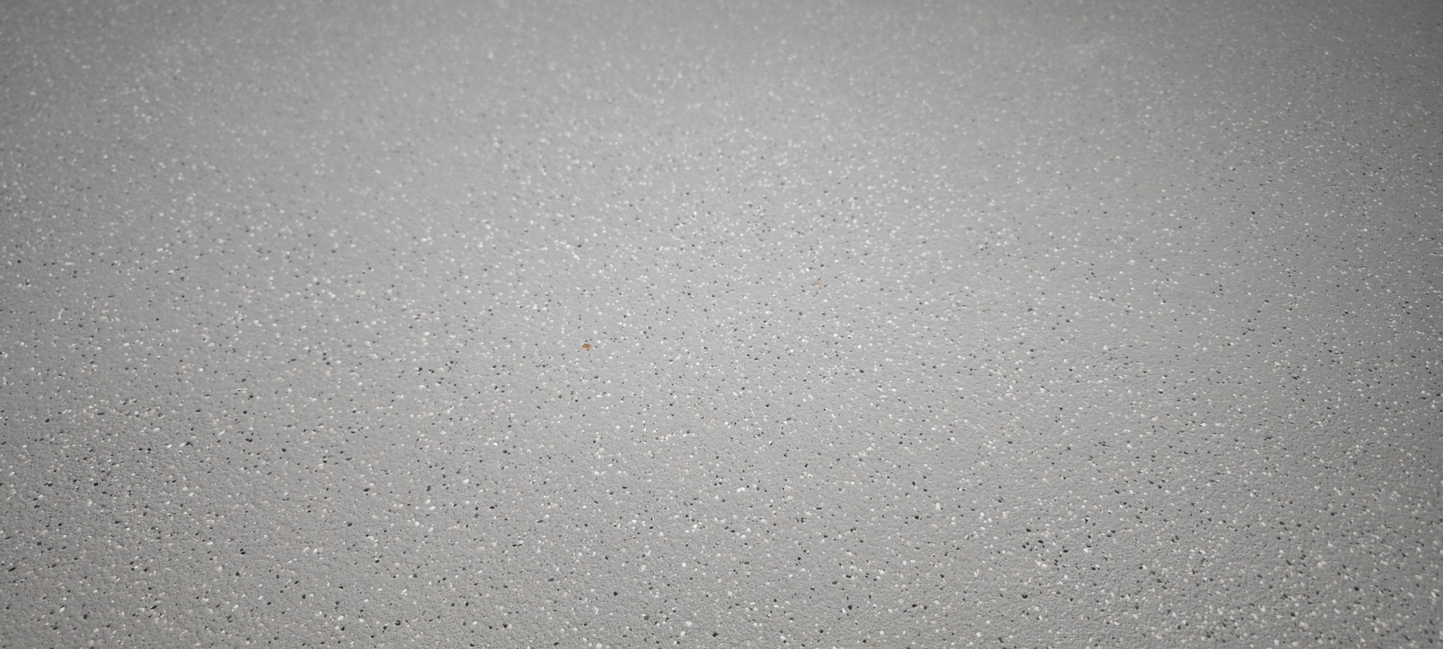Szara, gładka powierzchnia z drobnymi, ciemnymi i jasnymi inkluzjami, przypominająca wykończenie podłogi w Opacz Mała. Minimalistyczny wzór.