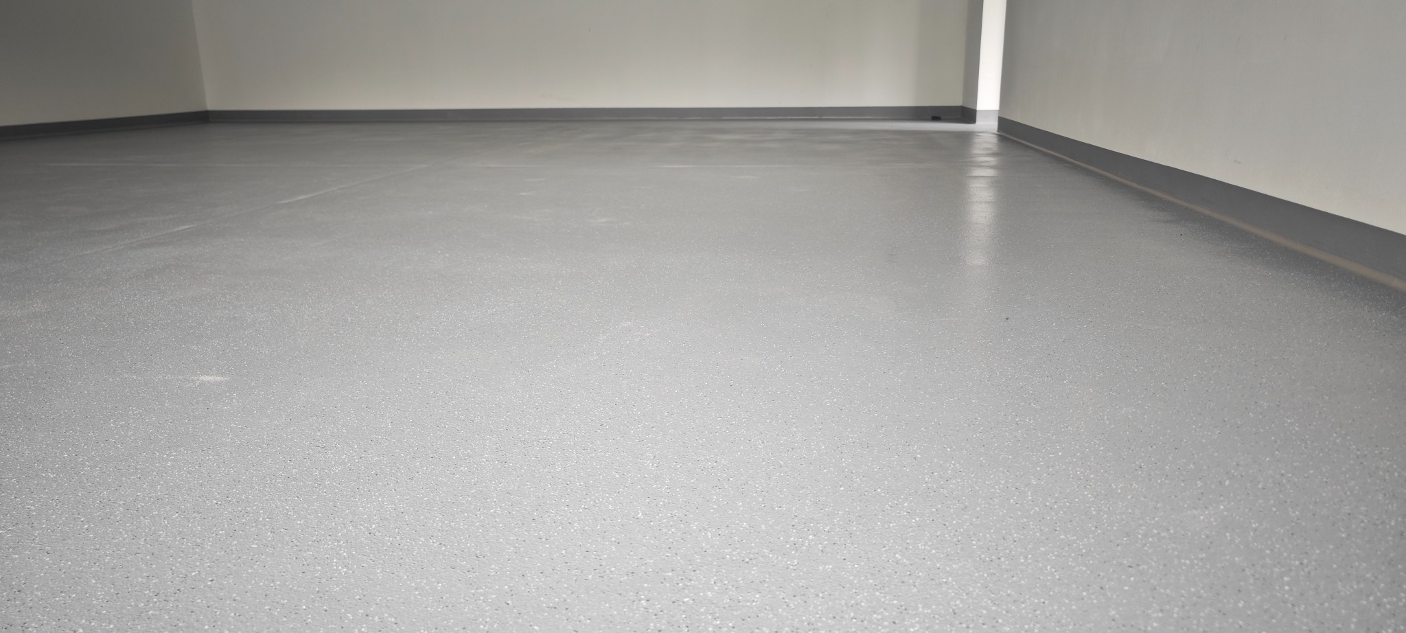 Jasnoszara, gładka posadzka z delikatnym wzorem, świeżo wylana w pustym pomieszczeniu. Widoczne listwy przypodłogowe i fragment ściany. Minimalistyczny design.