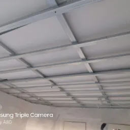 Metalowy stelaż sufitowy z zamontowanymi białymi płytami, widok z dołu, na płycie widoczny napis '47,5', przewody elektryczne zwisają z konstrukcji.