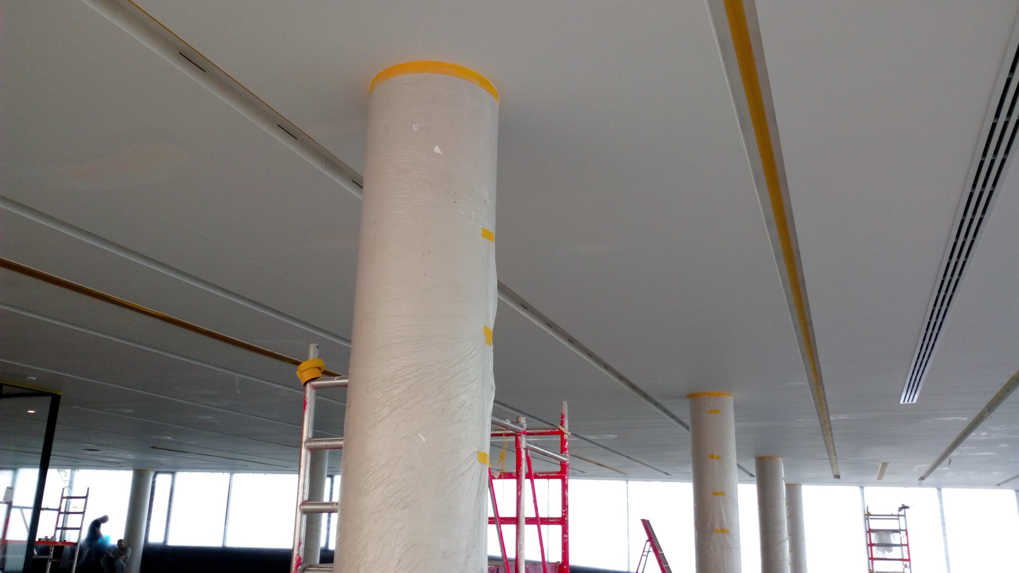 Przygotowanie do malowania sufitu w biurze: kolumny owinięte folią ochronną, taśma malarska na suficie, widoczne drabiny i pracownicy w tle.