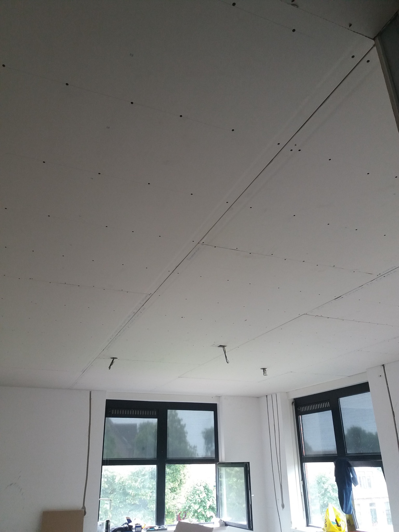 Widok sufitu z zamontowanymi płytami, widoczne ślady po wkrętach i łączenia płyt, okna z widokiem na zieleń w tle.