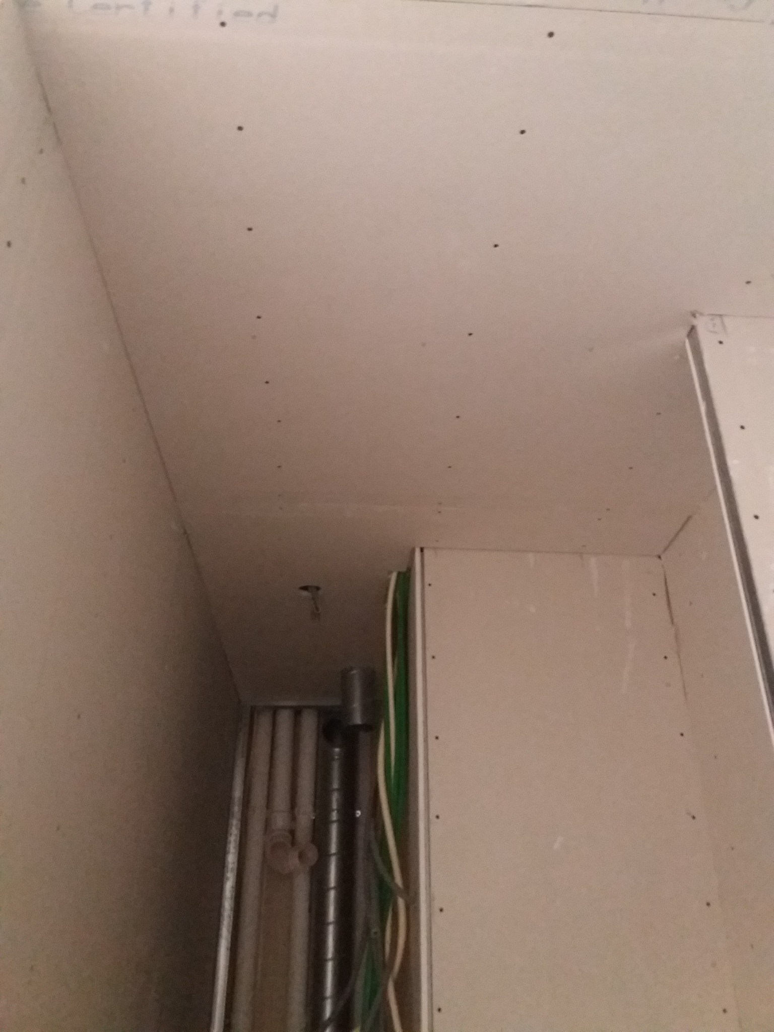 Narożnik pomieszczenia w trakcie prac, widoczne płyty gipsowo-kartonowe na ścianach i suficie, instalacje elektryczne i hydrauliczne w przestrzeni podsufitowej.