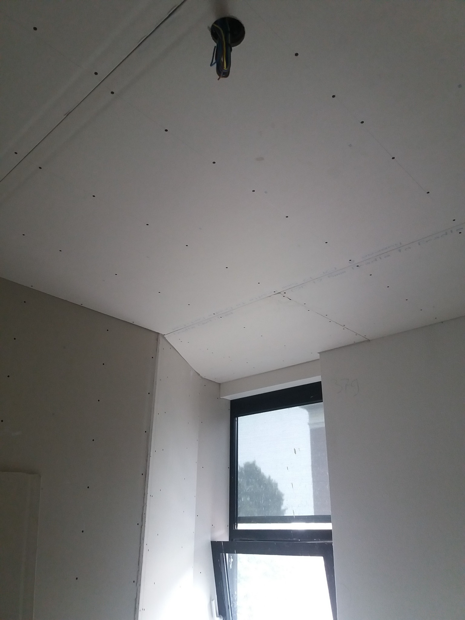 Montaż płyty karton gips na suficie i ścianach w pokoju z oknem, widoczne ślady po wkrętach, wystające kable ze stropu.