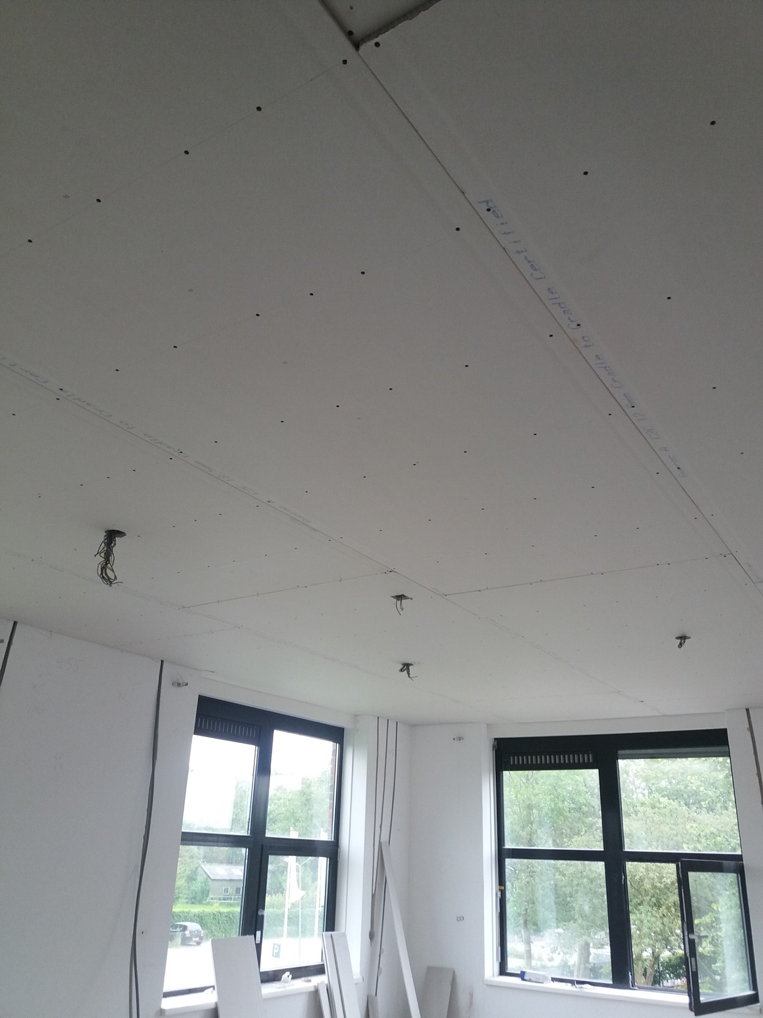 Montaż sufitu podwieszanego z płyt gipsowo-kartonowych w pomieszczeniu z dwoma oknami, widoczne kable wystające z otworów, ślady po szpachlowaniu i napisy na płytach.