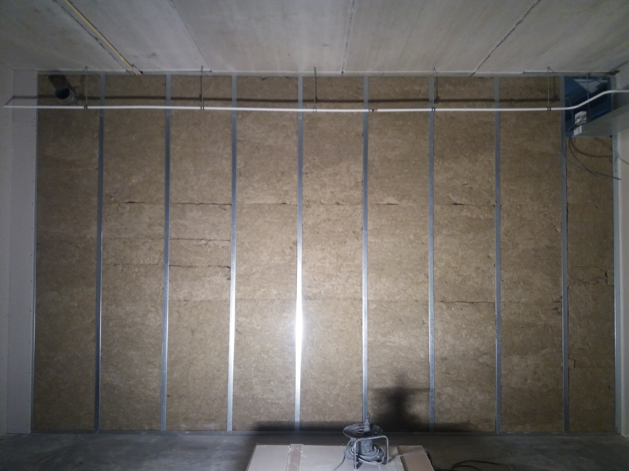 Ściana z izolacją z wełny mineralnej, zamontowana na metalowym ruszcie, widoczny przewód wentylacyjny pod sufitem i zwinięty kabel na podłodze.