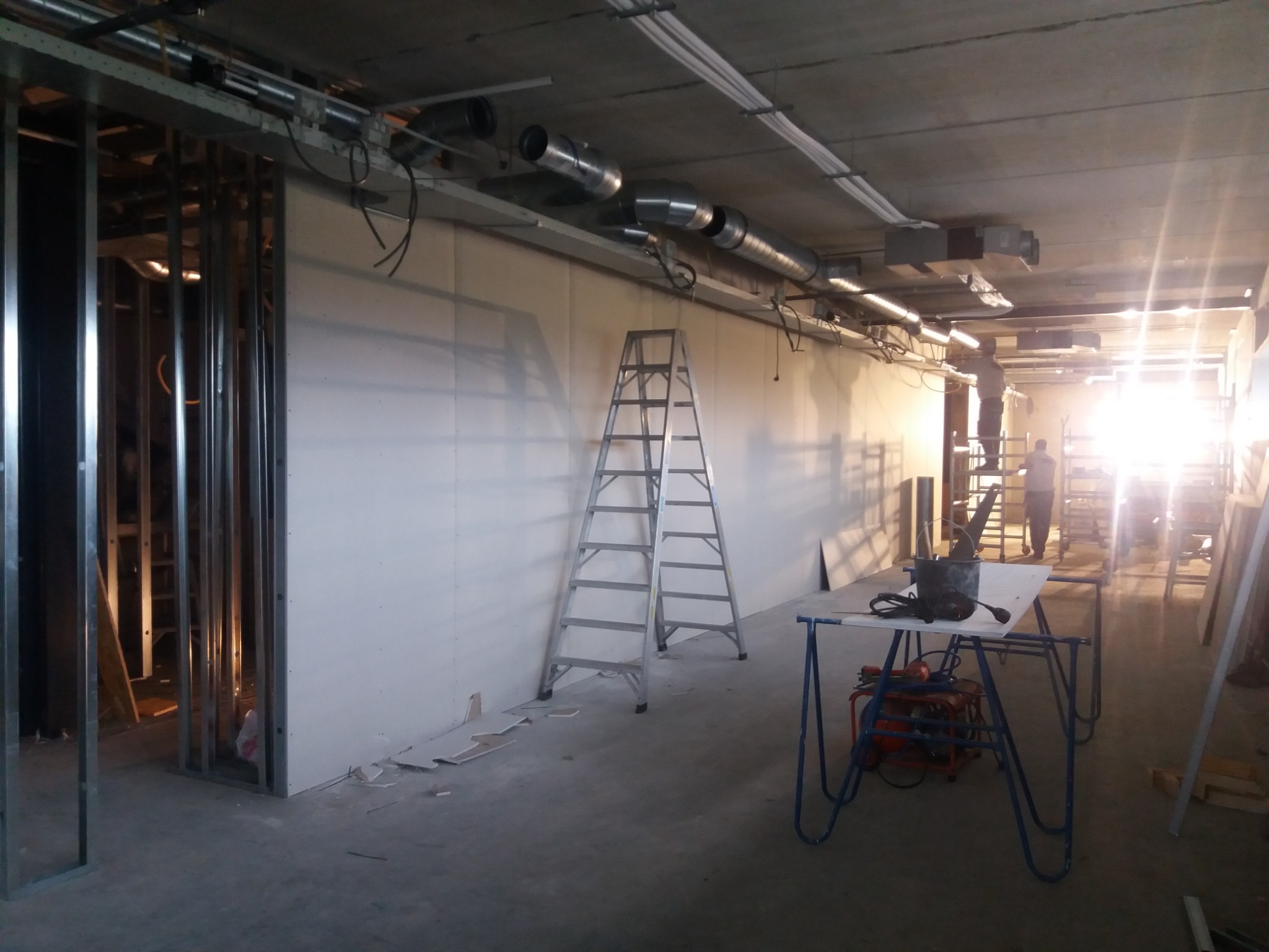 Wnętrze w trakcie prac wykończeniowych: metalowa konstrukcja ścian działowych, częściowo zamontowane płyty, drabina aluminiowa, instalacja wentylacyjna pod sufitem, narzędzia na stole warsztatowym.