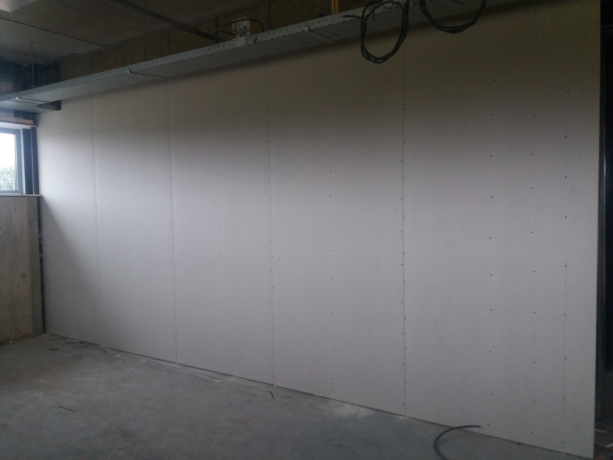 Ściana z płyt gipsowo-kartonowych, widoczne łączenia i wkręty, surowy betonowy sufit z instalacjami, okno w lewym górnym rogu, szary betonowy podkład podłogowy.