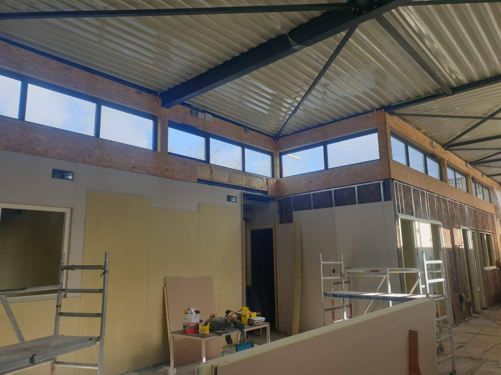 Wnętrze hali w trakcie prac wykończeniowych, widoczne ściany z płyt, konstrukcja dachu, okna pod sufitem oraz narzędzia na stole.