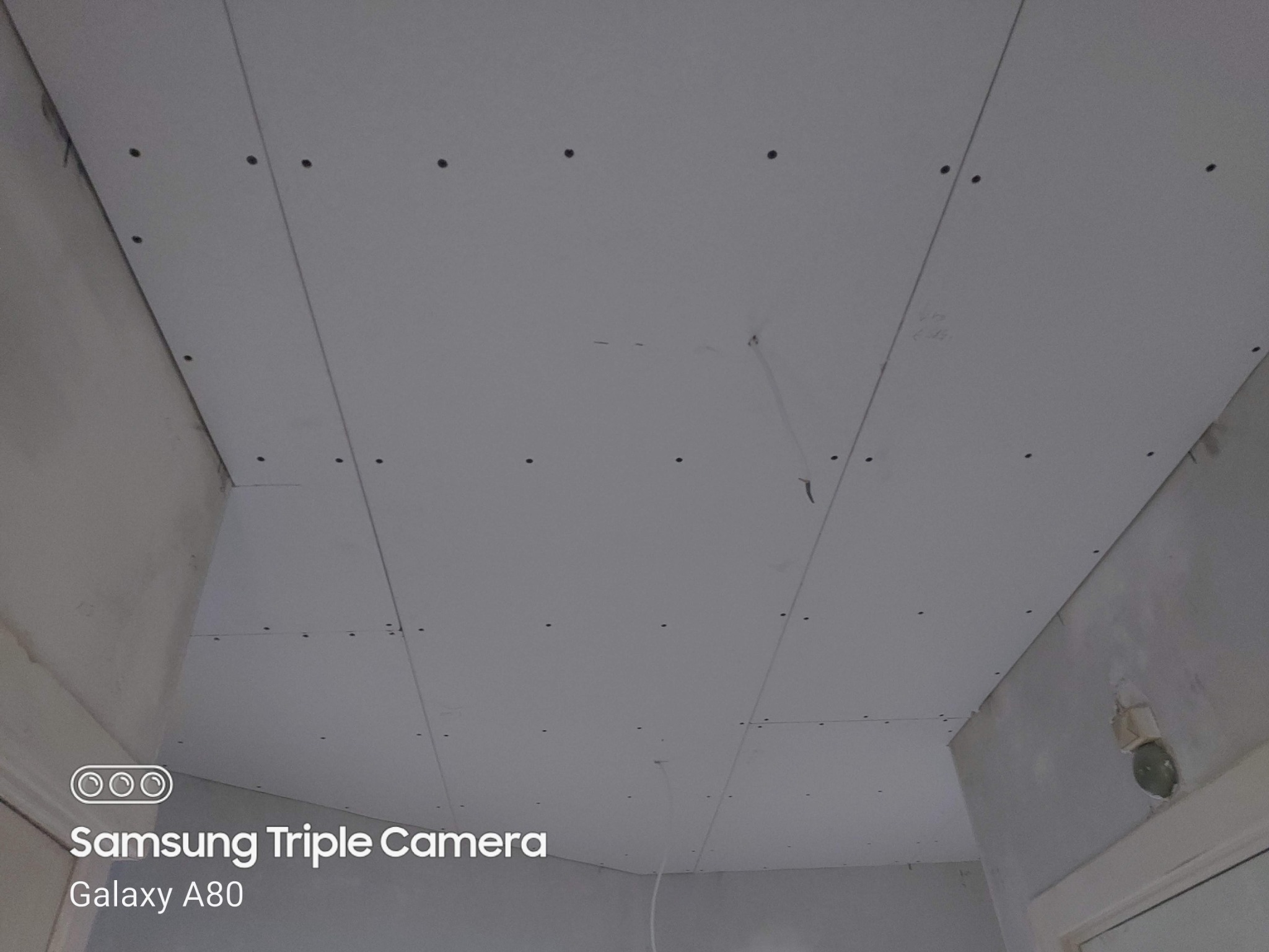 Sufit z zamontowanymi płytami gipsowo-kartonowymi, widoczne ślady po wkrętach, surowe wykończenie, fragment wystającego przewodu. Zdjęcie wykonane telefonem Samsung Galaxy A80.