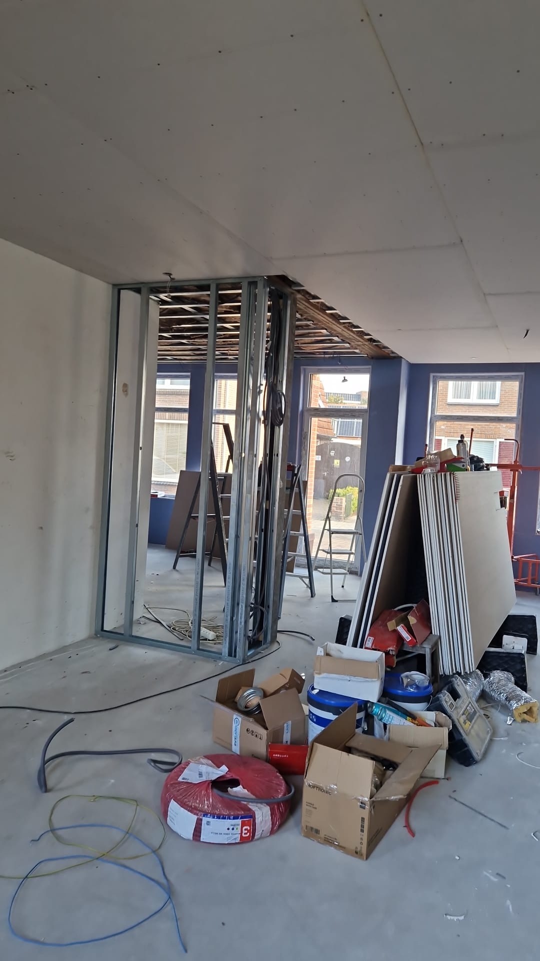 Wnętrze w trakcie prac: metalowy szkielet ścianki działowej, płyty, kable, drabiny. Widoczne okna i częściowo podwieszony sufit. Prace remontowe w jasnym pomieszczeniu.