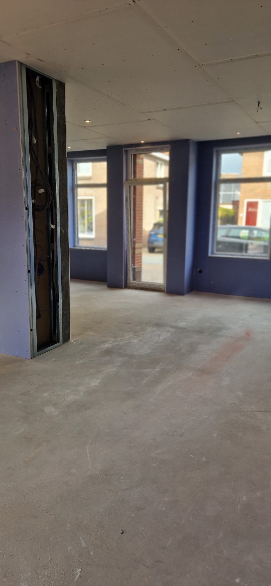 Wnętrze w trakcie prac: ściany w kolorze fioletowym, widoczne profile pod zabudowę, surowy beton na podłodze, okna z widokiem na zewnątrz. Etap przygotowania do dalszych prac.