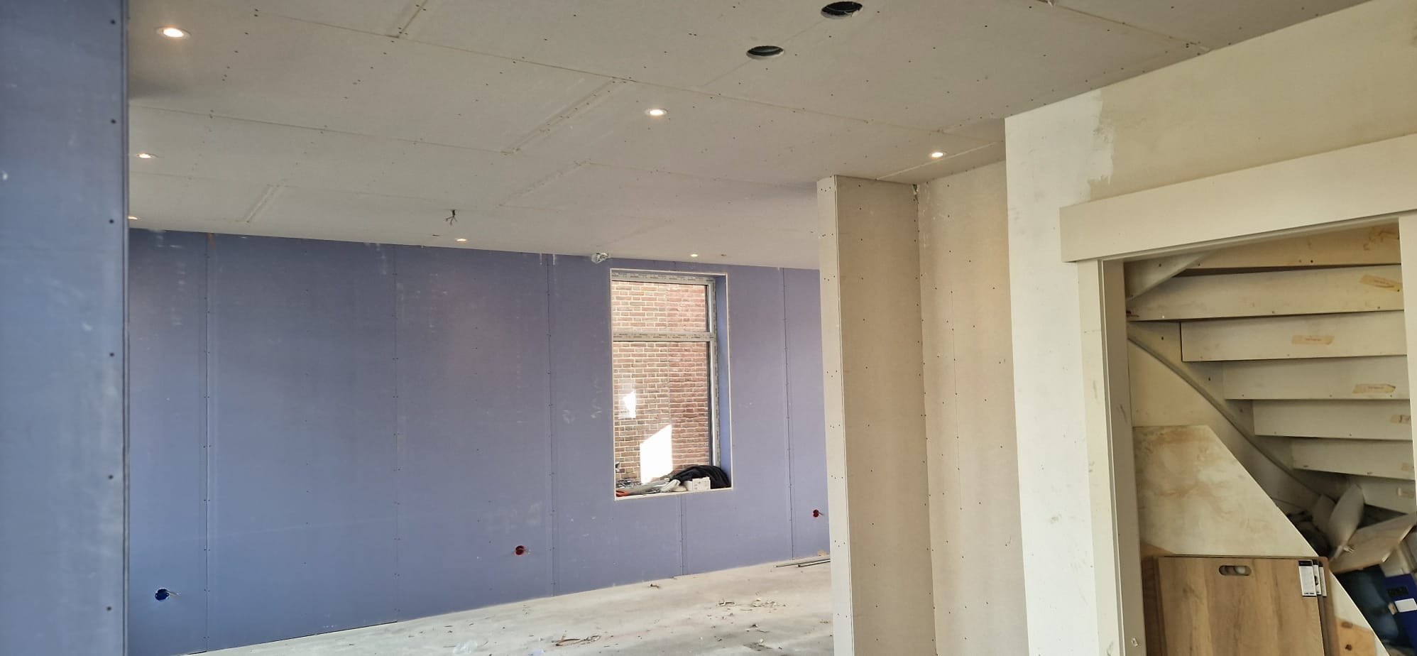 Wnętrze w trakcie prac: ściany obłożone płytami, widoczne punkty oświetleniowe w suficie, okno z widokiem na ceglany budynek, fragment schodów.