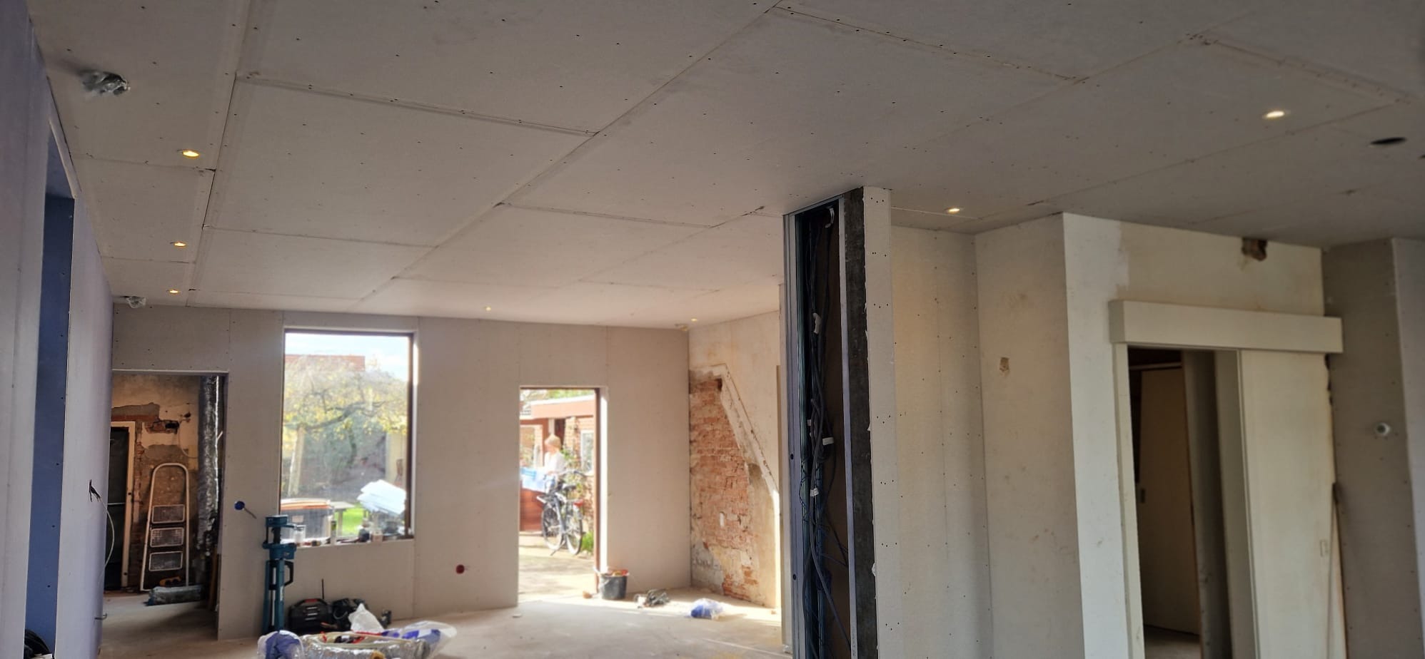 Wnętrze w trakcie prac wykończeniowych: zamontowane płyty na suficie z punktowym oświetleniem, ściany przygotowane do dalszych prac. Widoczne okna i instalacje.