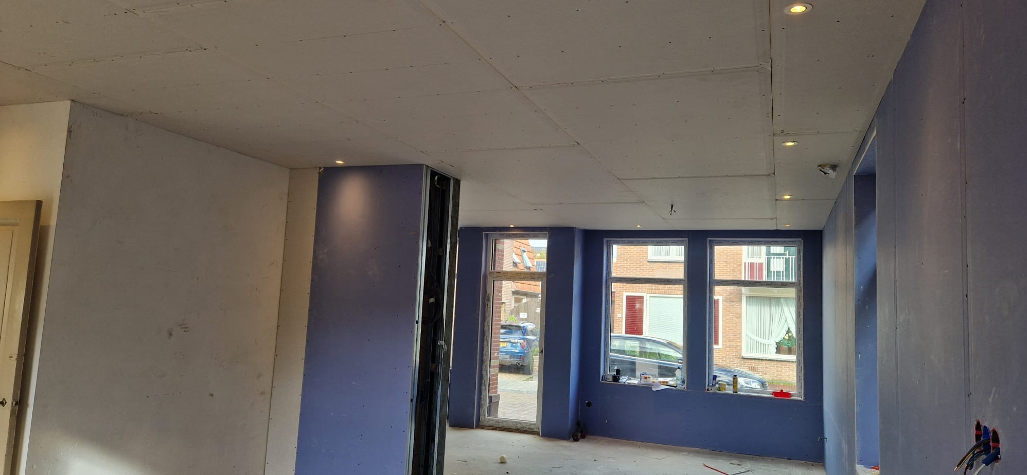 Wnętrze w trakcie remontu: ściany z płyt karton-gips w odcieniach fioletu i szarości, sufit z zamontowanym oświetleniem punktowym, okna z widokiem na zabudowę miejską.
