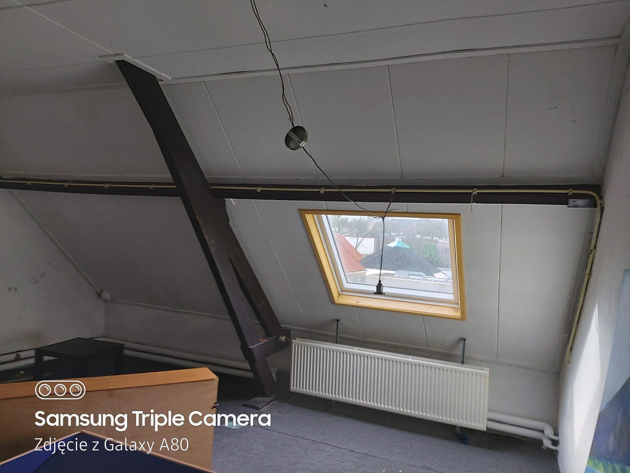 Poddasze ze skośnym sufitem wykończonym panelami, widoczne okno dachowe, grzejnik i elementy instalacji elektrycznej. Ukończone prace wykończeniowe.