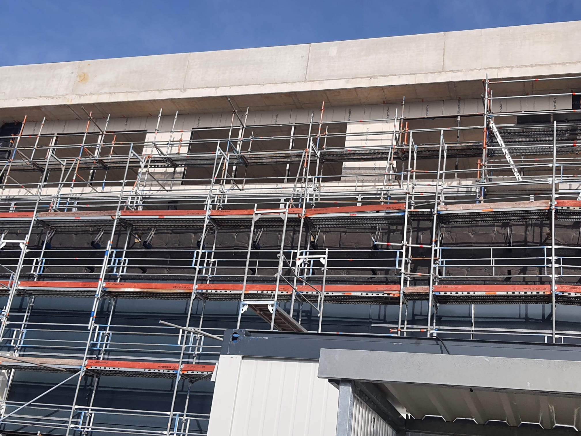 Budynek w trakcie prac wykończeniowych, widoczne rusztowania i zabezpieczenia. Jasne niebo w tle. Ujęcie z dołu, podkreślające wysokość konstrukcji.