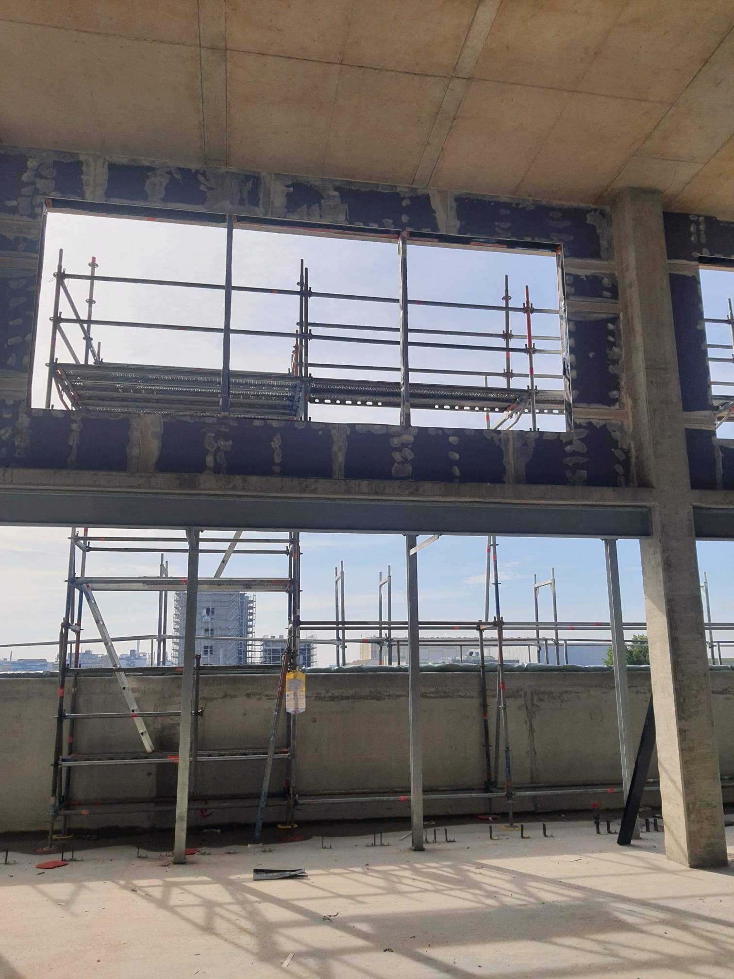 Wnętrze w surowym stanie z widocznymi rusztowaniami i przygotowanymi otworami okiennymi. Widoczny betonowy strop i słupy konstrukcyjne. Prace wykończeniowe w toku.