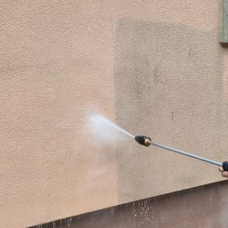 MultiFach - Czyszczenie elewacji budynku myjką ciśnieniową. Widoczna różnica w kolorze między umytą a brudną częścią ściany. Woda spływa po elewacji i kostce brukowej.